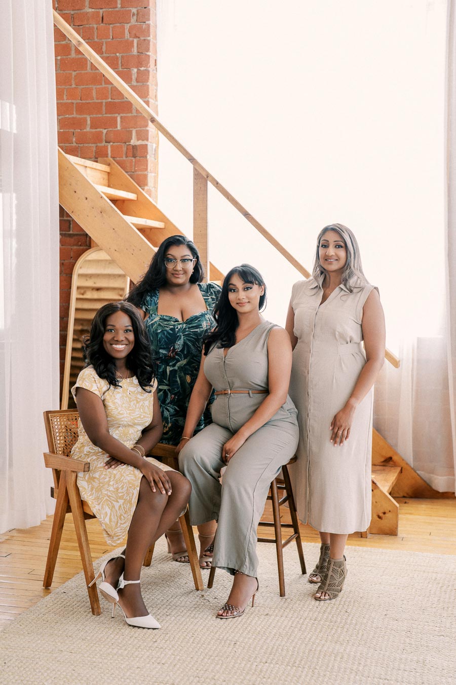 Four women pose together in a bright room with a wooden staircase, showcasing a mix of stylish outfits.