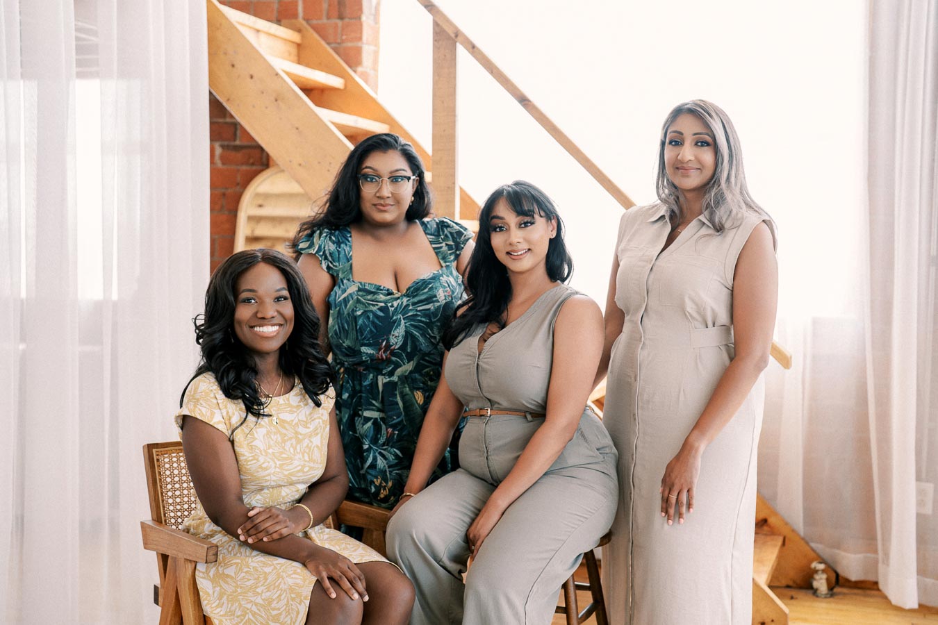A group of four women posing together indoors, with a wooden staircase in the background. They are dressed in stylish outfits, smiling and looking at the camera.