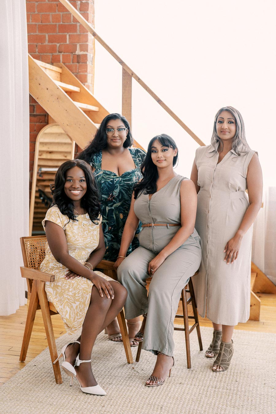 Four women pose together in a stylish indoor setting, featuring wooden stairs and a brick wall.