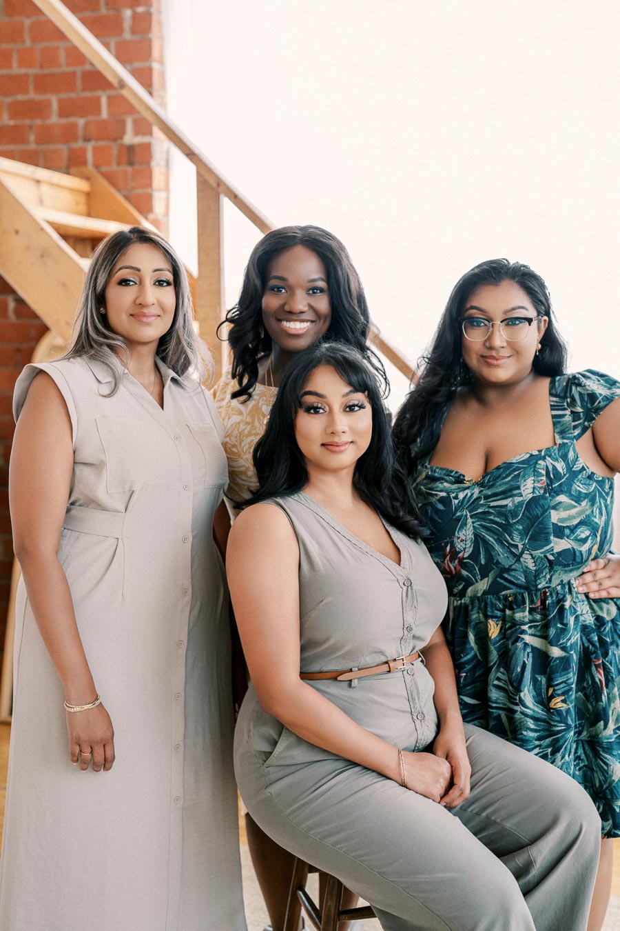 A group of four women smiling and posing together indoors, with a brick wall and wooden staircase in the background.