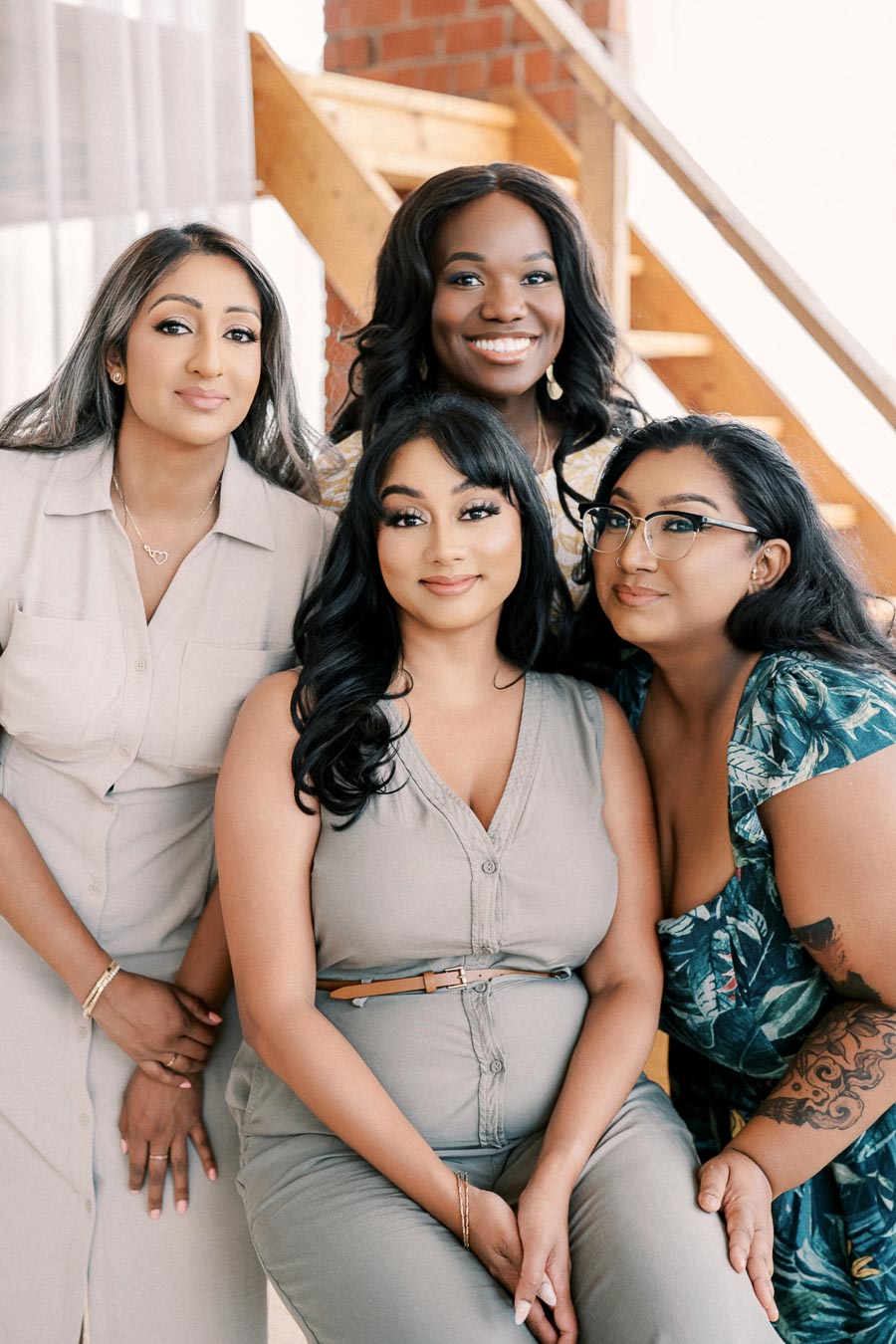 Four women smiling and posing together indoors, with a staircase in the background.
