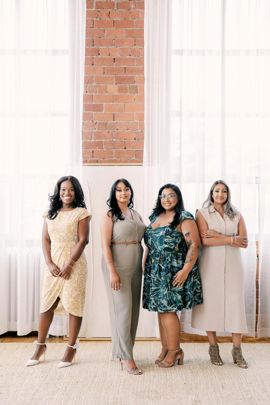 Four women pose confidently in front of a brick wall, each wearing a different stylish dress. The setting is bright and airy, with natural light streaming through large windows.