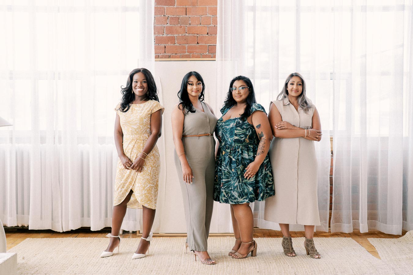 Four women pose confidently in a bright, stylish room, showcasing diverse fashion choices.