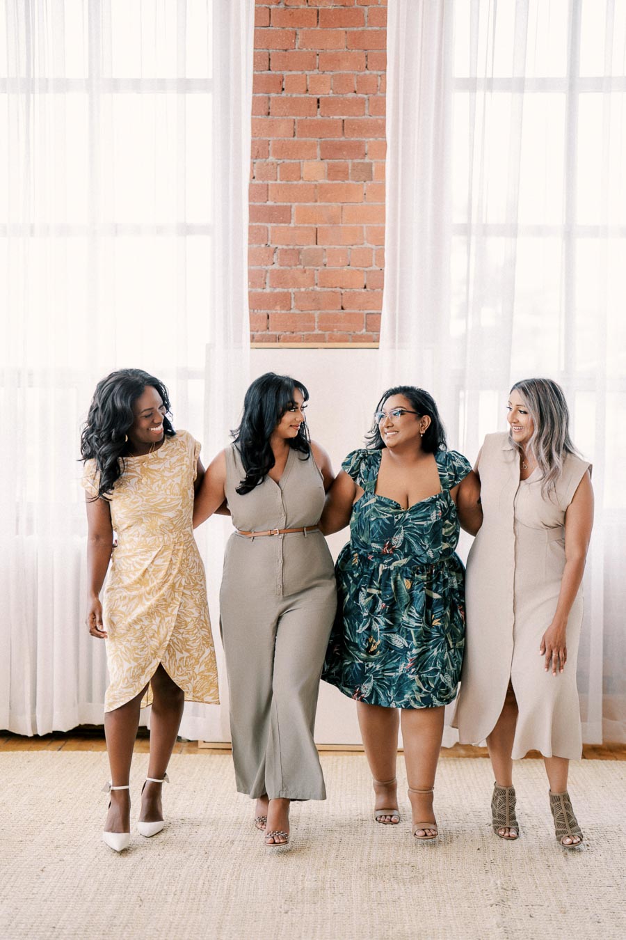 Four women stand together in a warmly lit room with brick walls and white curtains, each wearing stylish dresses and smiling at each other.