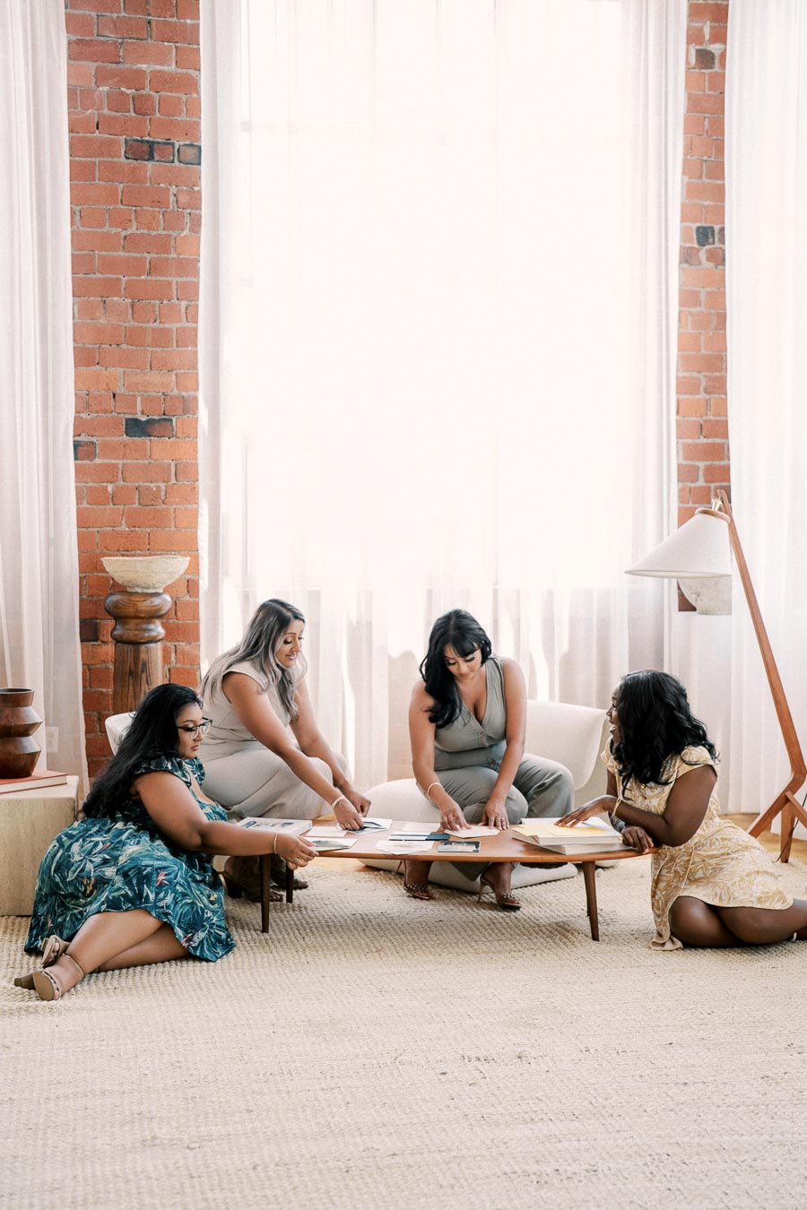 Four women engage in a collaborative discussion around a low wooden table, surrounded by natural light and brick walls, creating a cozy and inviting atmosphere.