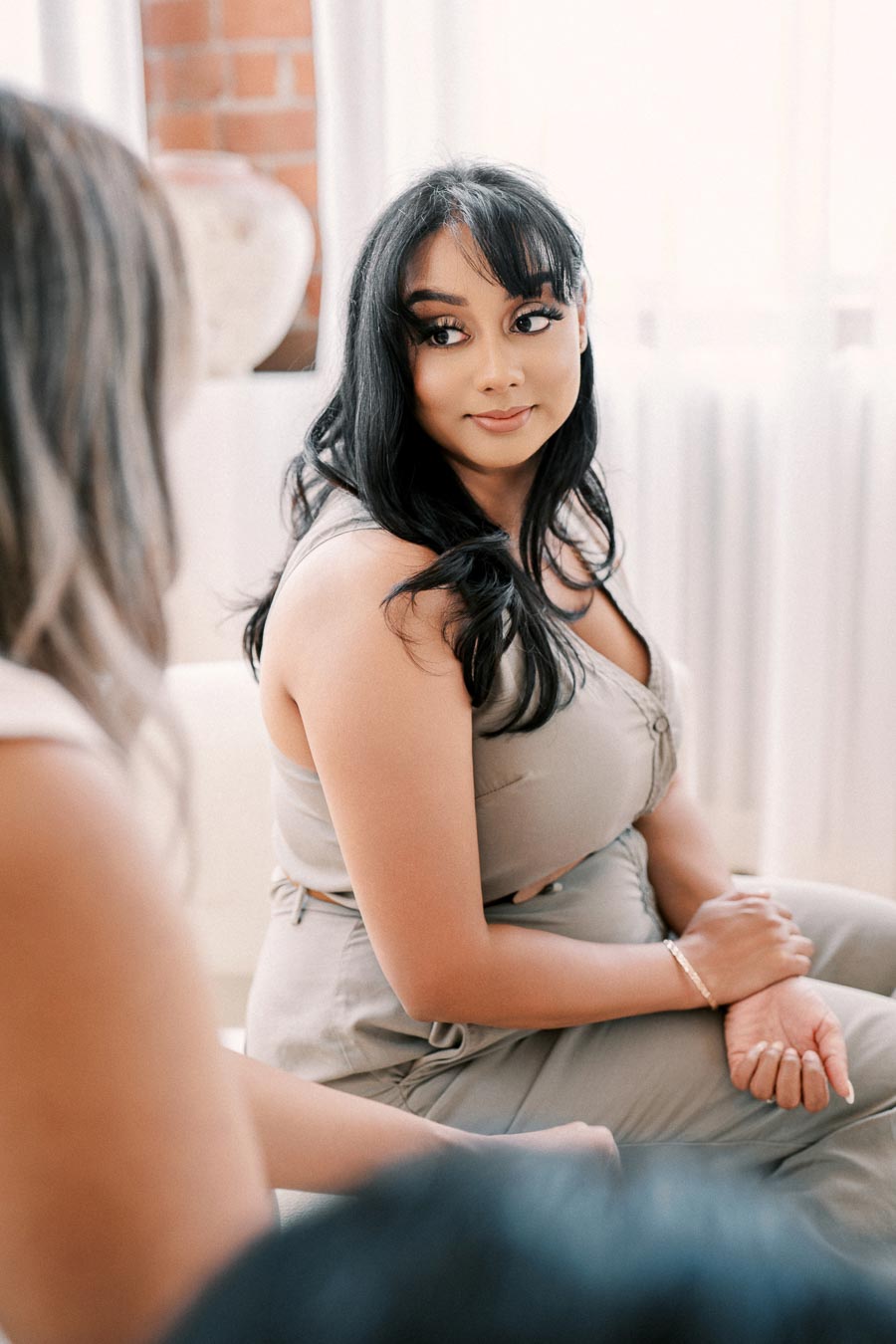 A woman with long dark hair and bangs sits in a bright room, engaging in conversation.