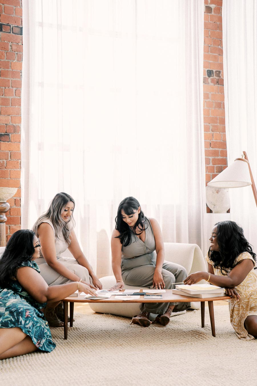 Four women gather around a coffee table, engaged in conversation in a cozy room with brick walls and large windows.