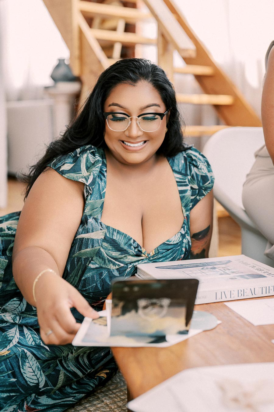 A woman in a leaf-patterned dress enjoys a moment at a table, surrounded by books and photographs.