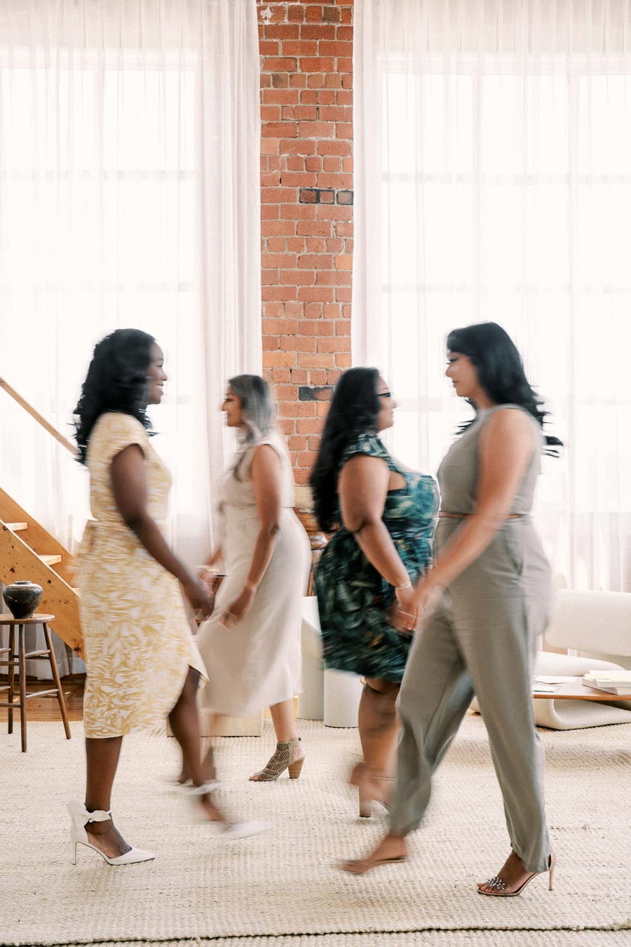 Four women walk gracefully in a stylish room with exposed brick walls and large windows, creating a sense of movement and elegance.