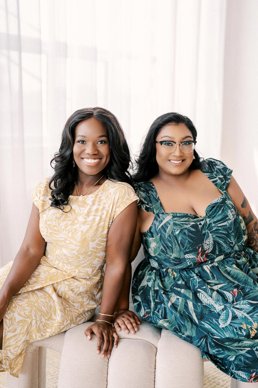 Two women in floral dresses smiling while sitting together in a bright room.