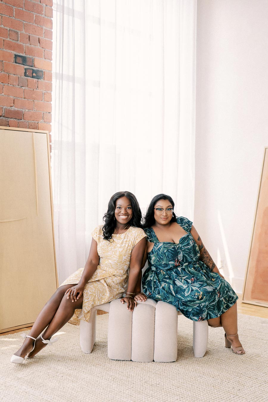 Two women in floral dresses sit on a cushioned bench in a bright, airy room with brick walls and large windows.