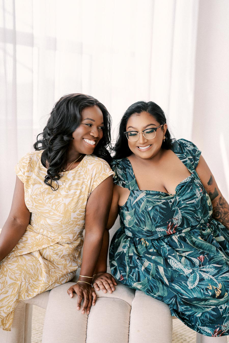 Two women in floral dresses share a joyful moment, sitting together and smiling.