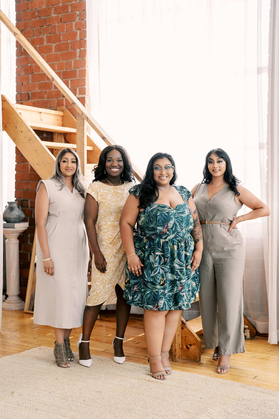Four women pose together indoors, standing in front of a wooden staircase and brick wall. They are dressed in stylish, summery outfits and smiling at the camera.