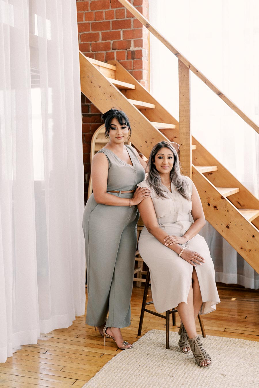 Two women pose elegantly in a stylish indoor setting with a wooden staircase and brick wall backdrop. Both are dressed in neutral-toned outfits, with one standing and the other seated on a stool.
