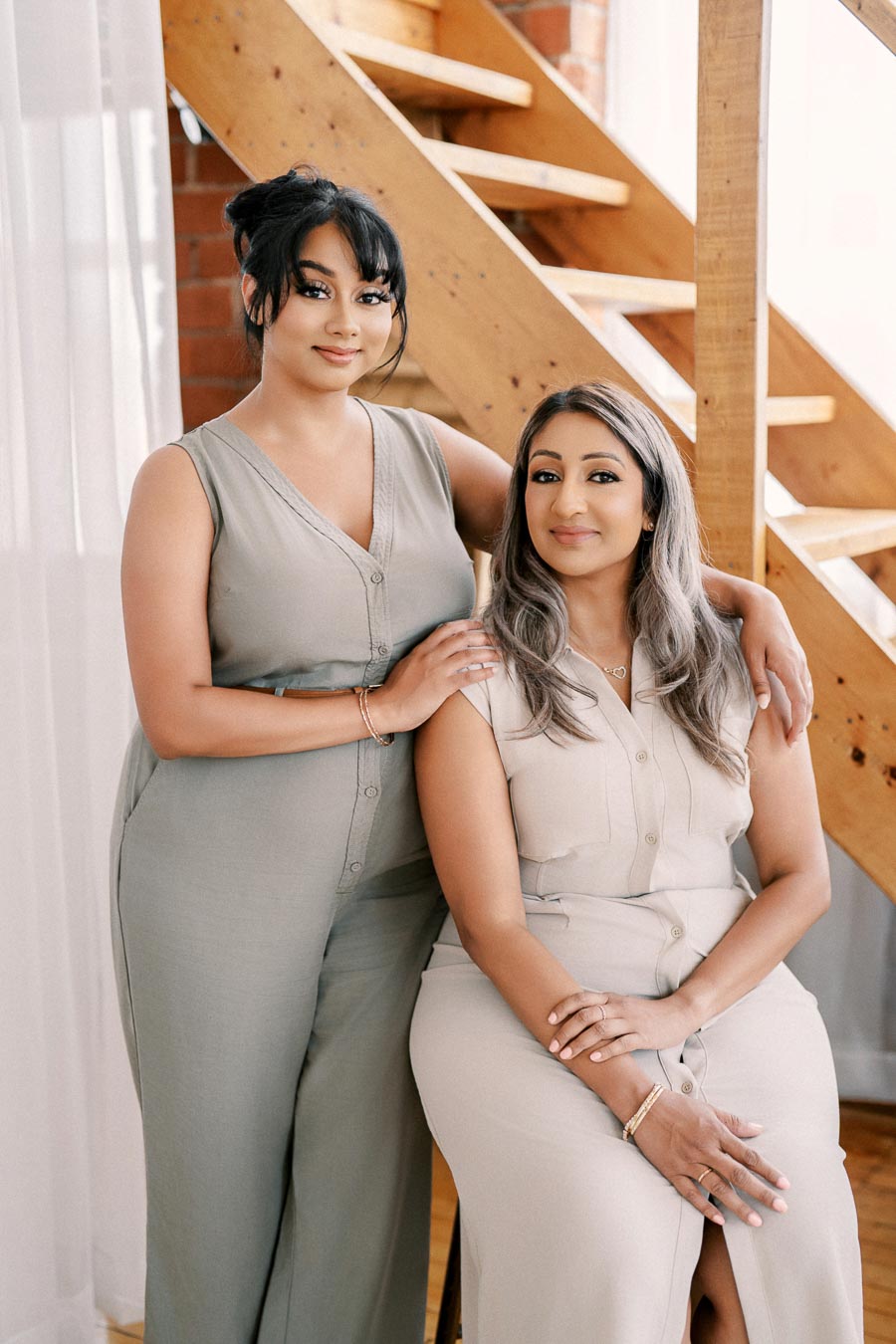 Two women pose together indoors, dressed in light-colored outfits, with a wooden staircase in the background.