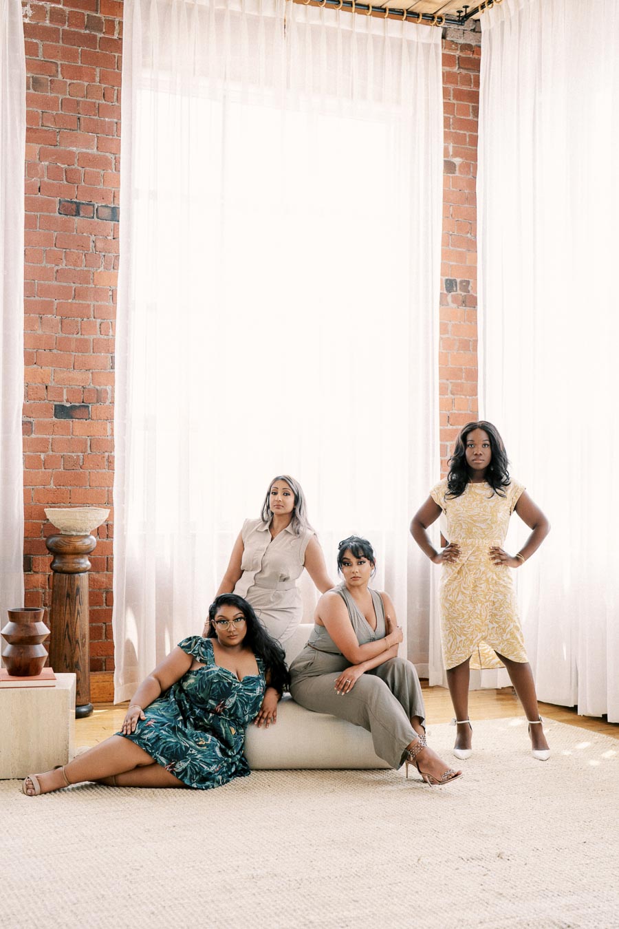 Four women pose confidently in a stylish room with exposed brick walls and large windows, dressed in elegant outfits.