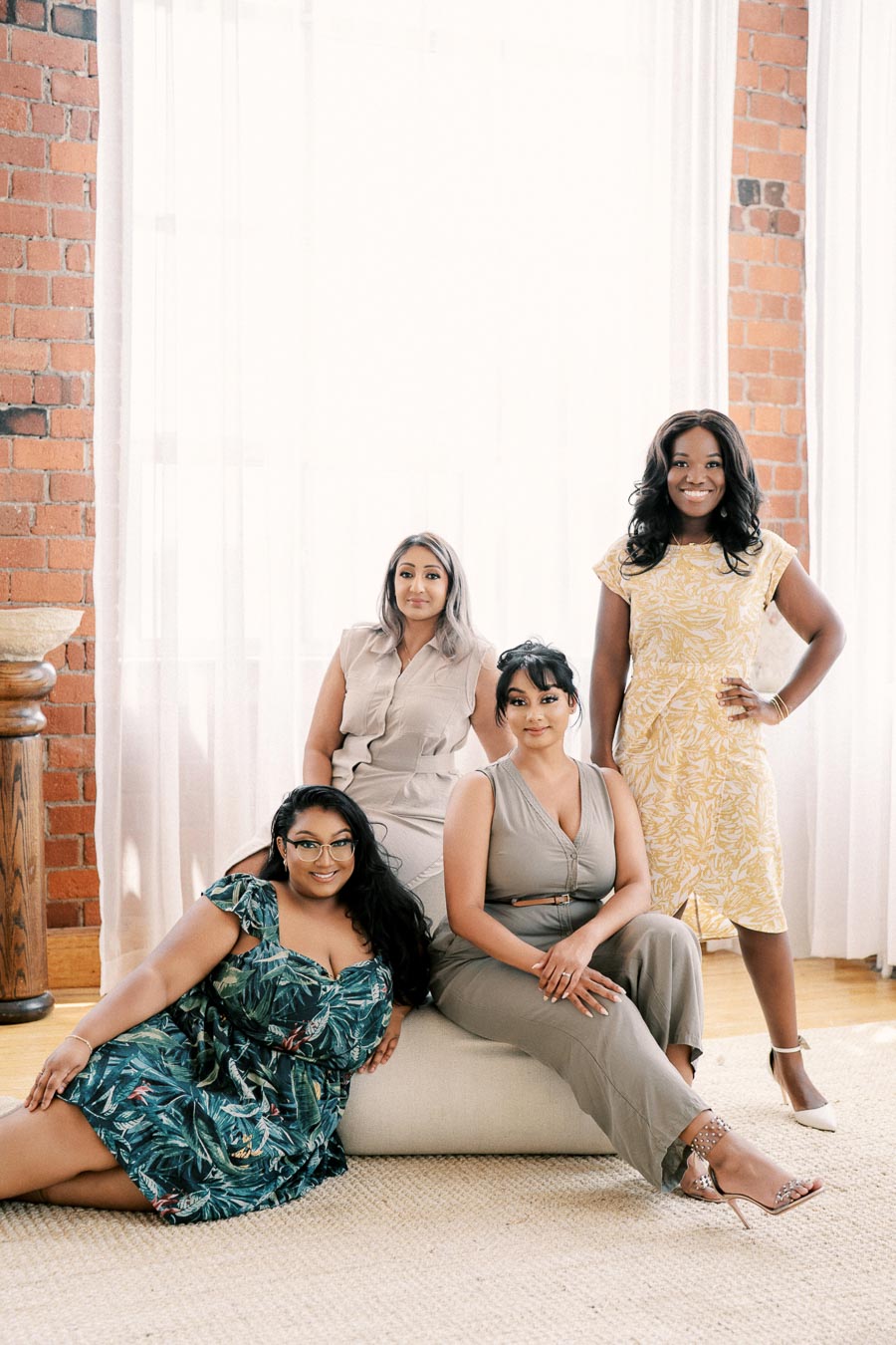 Four women pose together in a stylish room with brick walls and soft lighting, showcasing a variety of fashionable outfits.