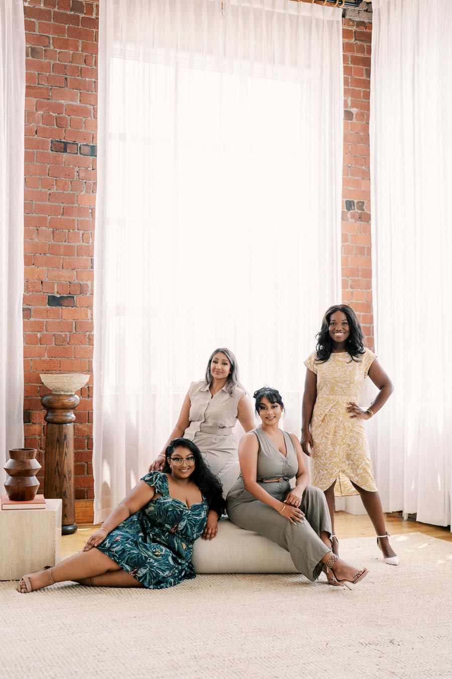 Four women pose together in a well-lit room with brick walls and sheer curtains, showcasing a stylish and relaxed atmosphere.