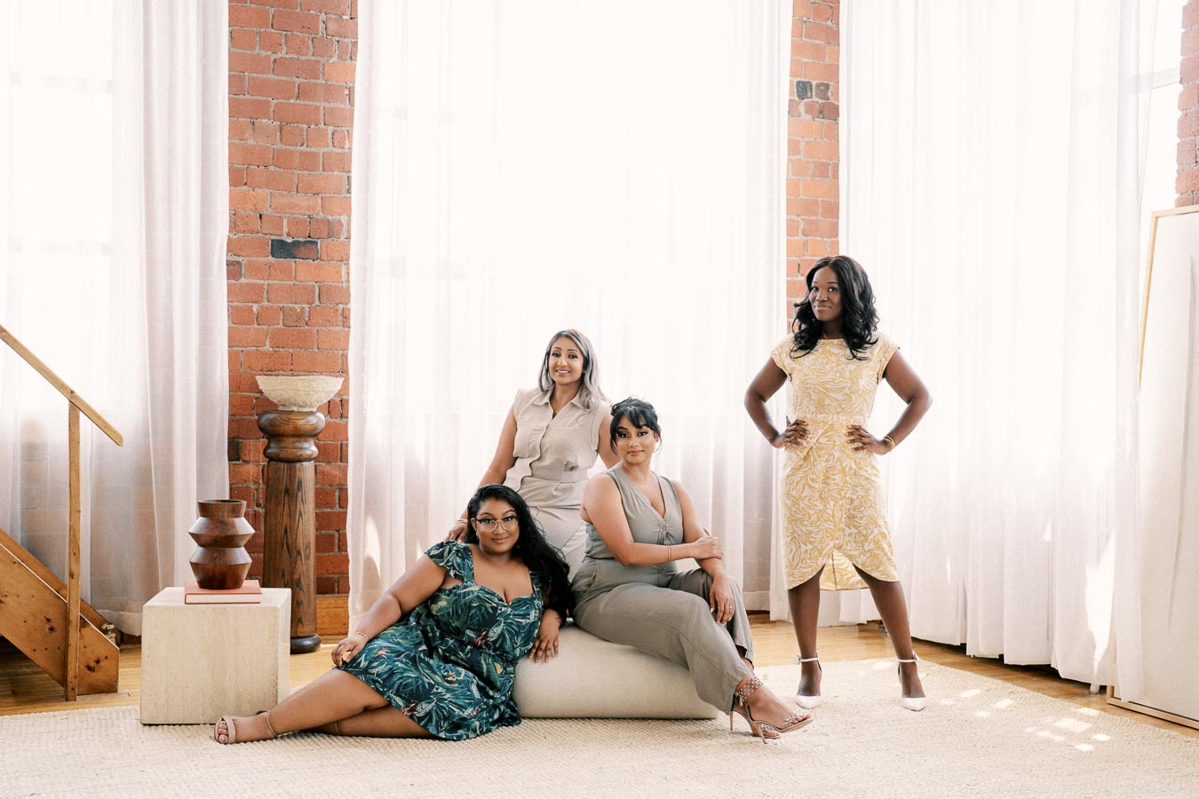 Four women pose confidently in a well-lit room with brick walls and sheer white curtains, showcasing a mix of elegant and casual attire.
