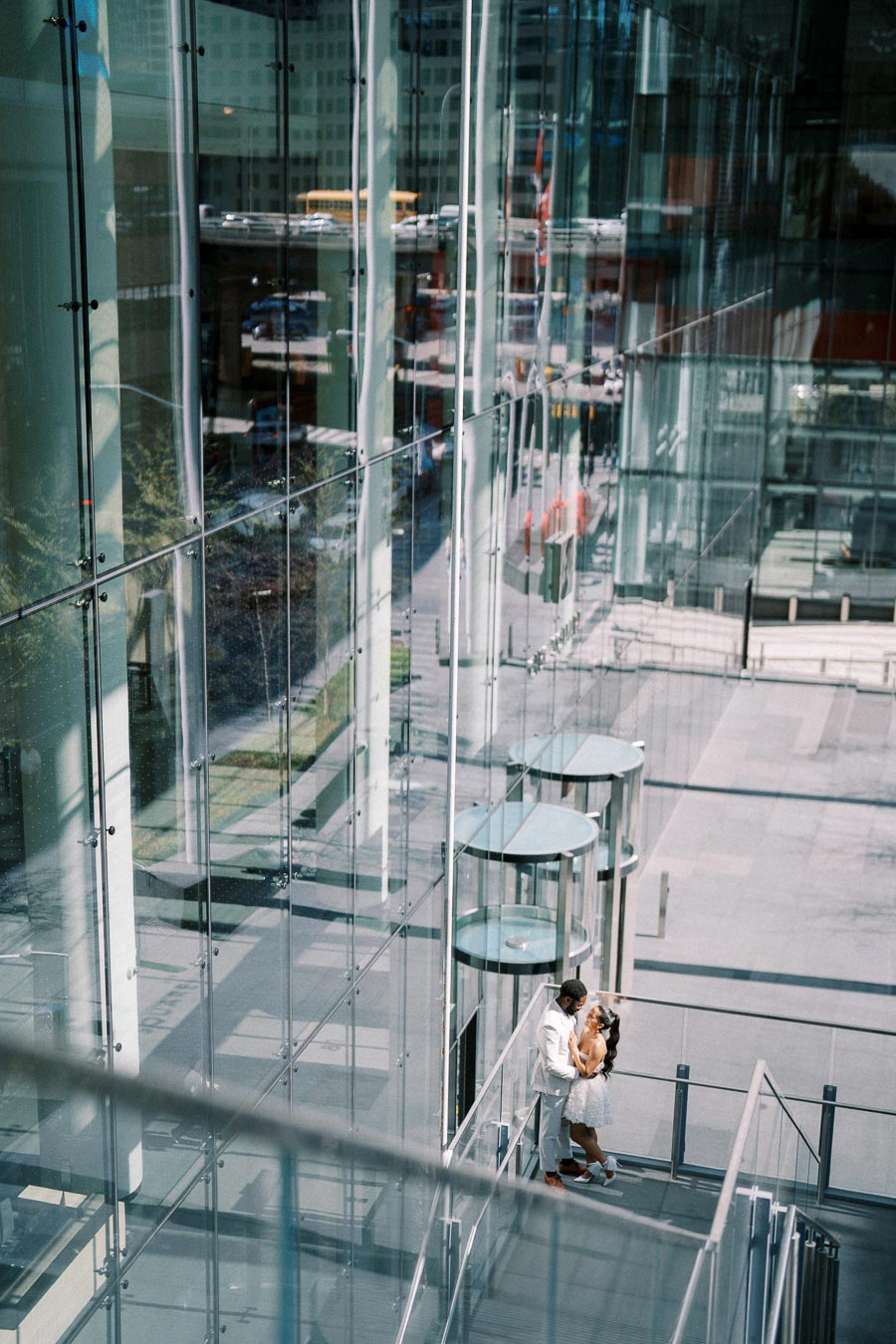 A couple embraces on a modern glass balcony, surrounded by urban architecture with reflections of city life visible on the windows. The woman is in a white dress and the man is in a light-colored suit.