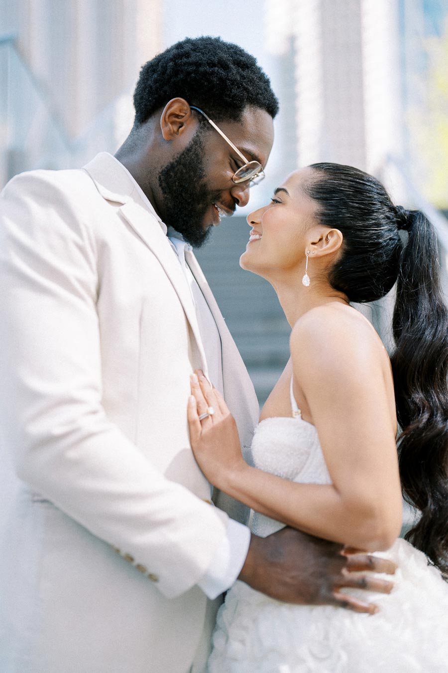 A joyful couple in wedding attire embracing and smiling at each other outdoors, capturing a romantic moment. The bride wears a white dress and elegant earrings, while the groom is in a light-colored suit with glasses.