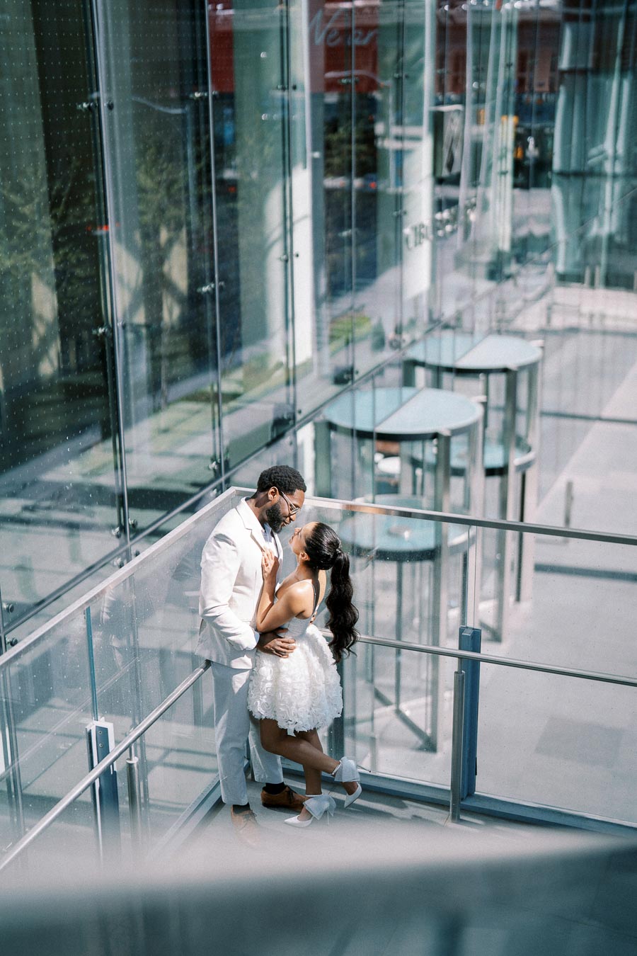 A romantic couple dressed in elegant attire embraces in a modern glass building with urban views.