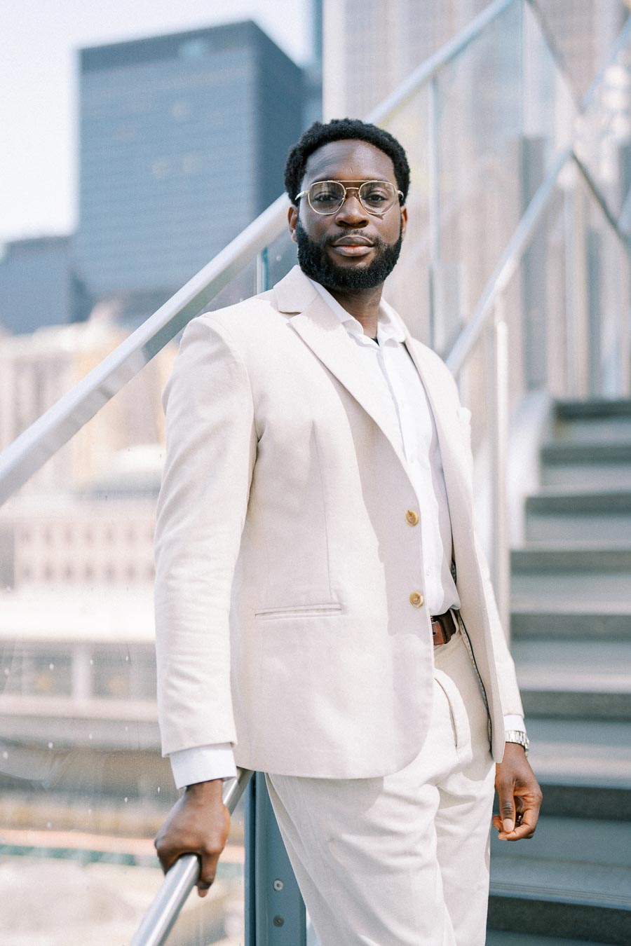 Portrait of a professional man in a light-colored suit standing on urban stairs, with modern skyscrapers in the background.