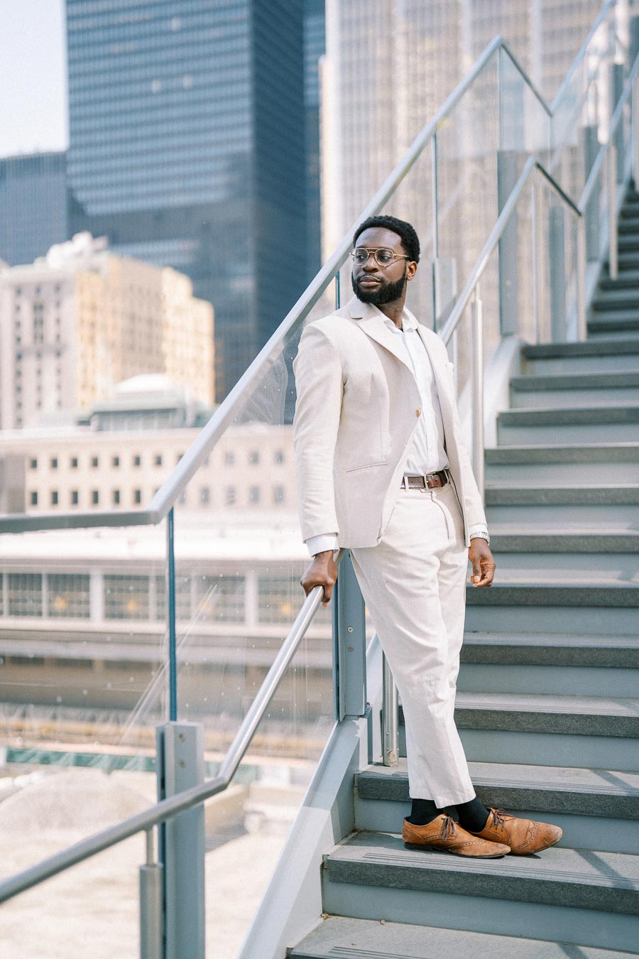 Man in a stylish white suit standing on outdoor stairs with a modern cityscape backdrop, exuding confidence and elegance.