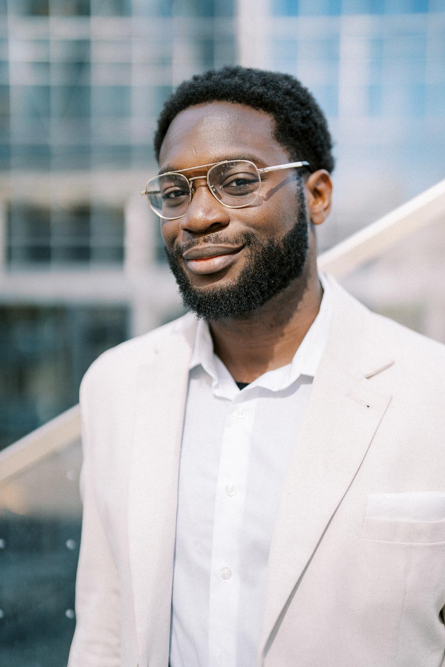 Stylish man with a beard and glasses in a white suit standing outside a modern office building.