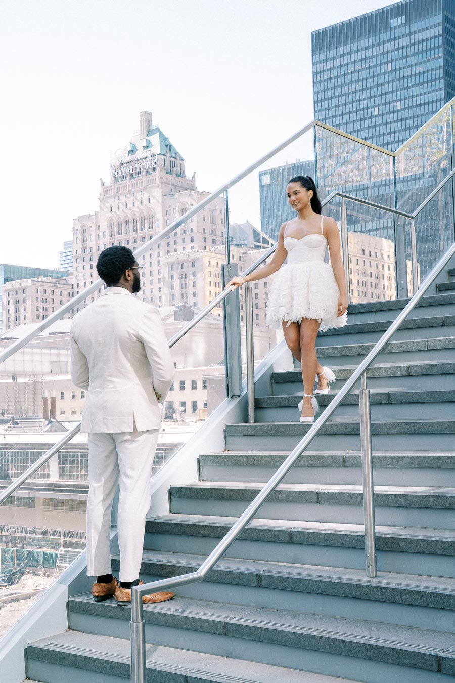 A couple in elegant attire stands on an outdoor staircase, with a cityscape of tall buildings in the background. The woman in a white dress descends the stairs towards the man in a light-colored suit, capturing a romantic urban moment.