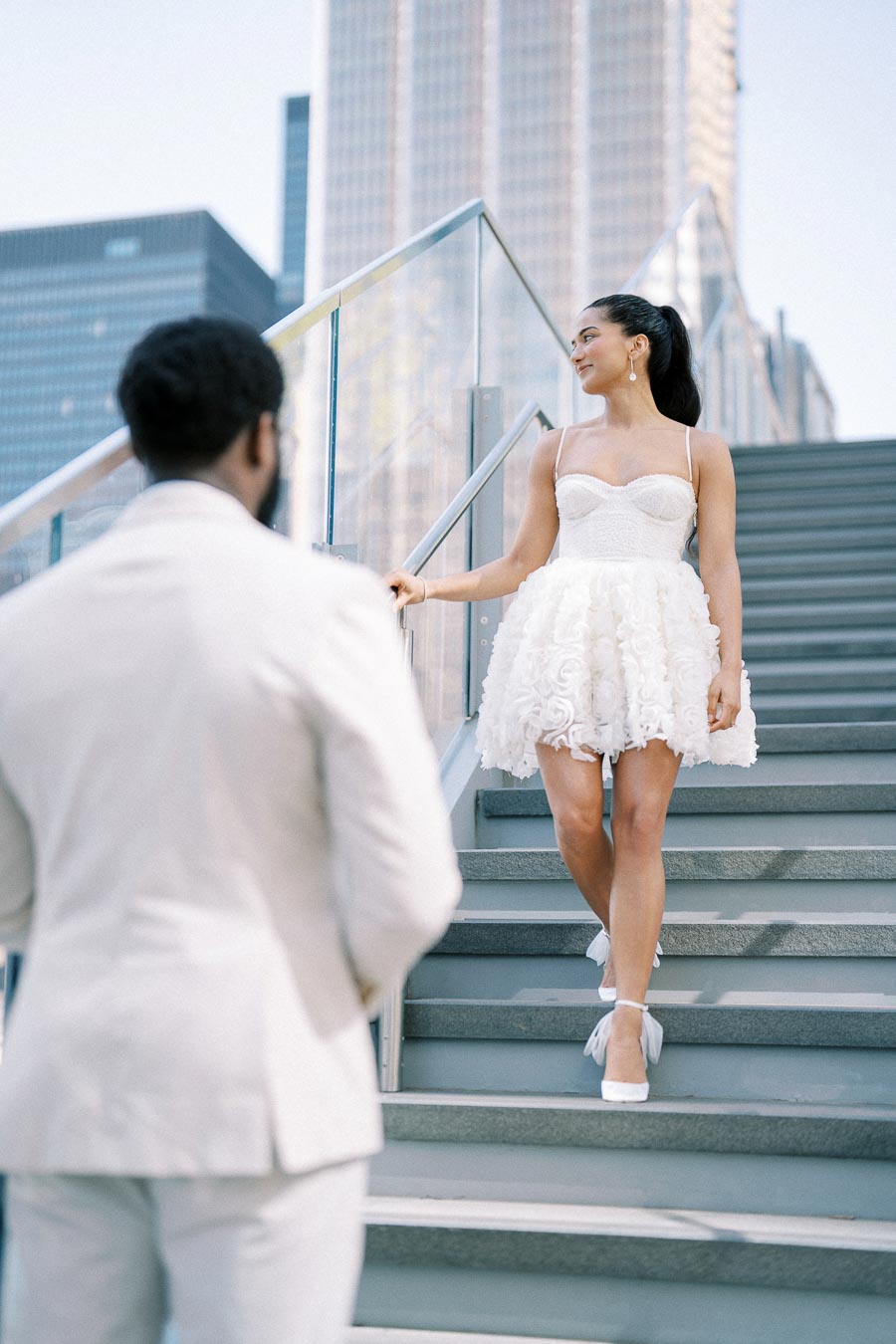 A woman in a white dress descends stairs outdoors, smiling at a man in a light-colored suit with a city skyline in the background.