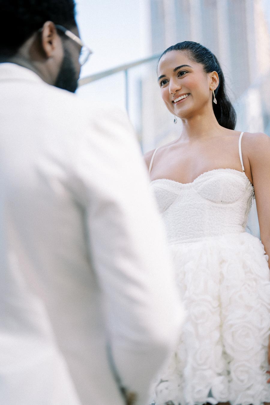 Bride smiling at groom during outdoor wedding ceremony, both dressed in elegant white attire.