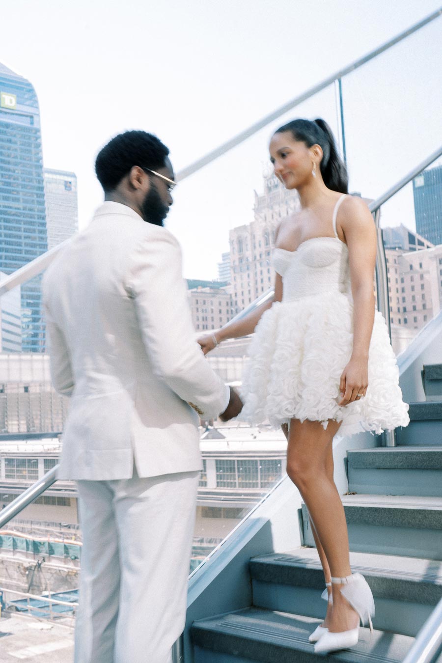 Elegant couple in white attire on a staircase, with a cityscape background, capturing a stylish urban moment.