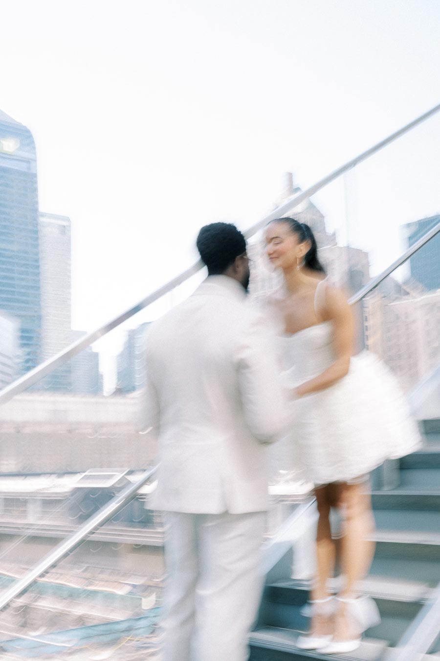 Blurred image of a couple on a staircase with cityscape background, man in white suit and woman in white dress.