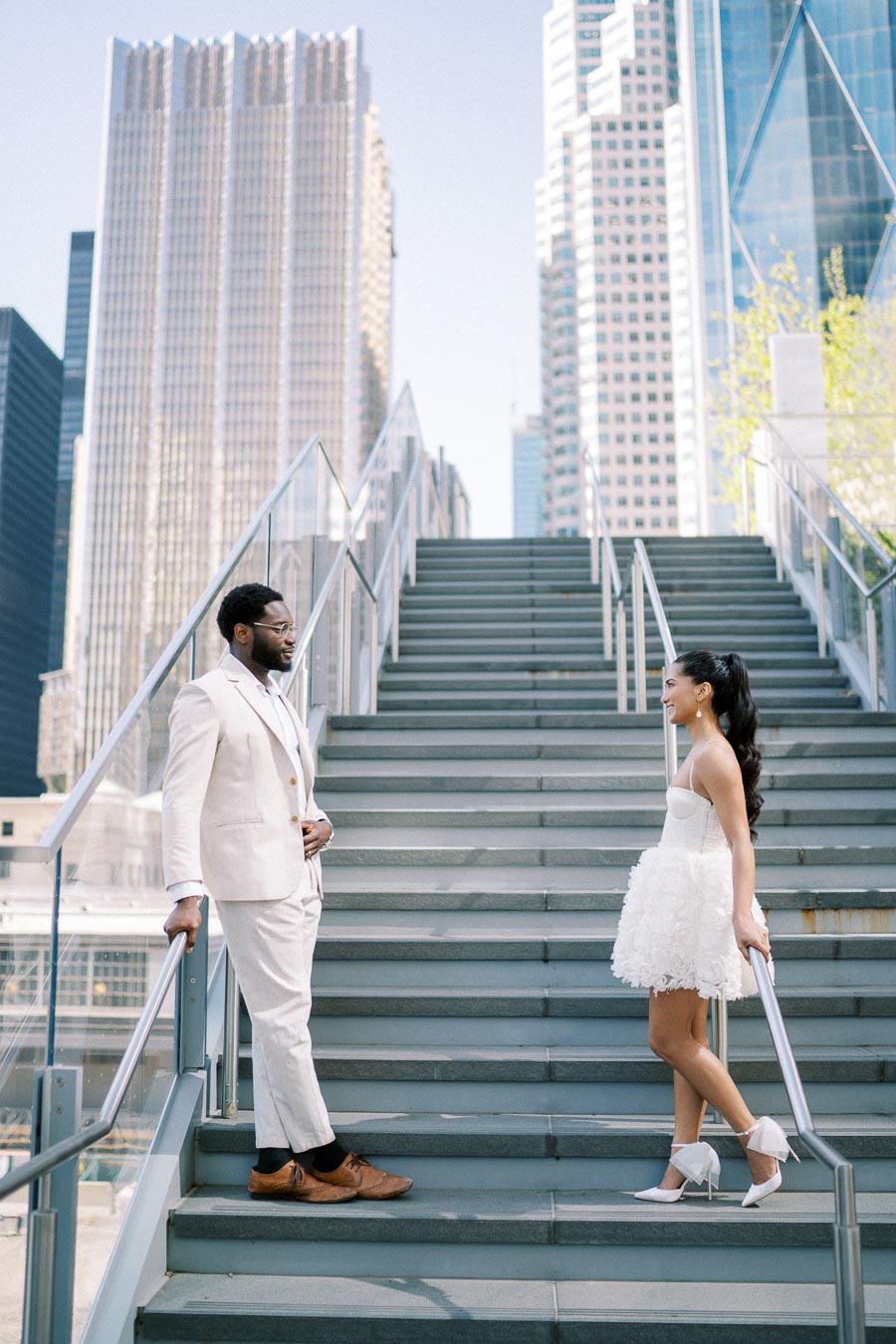 A couple dressed in elegant white attire stands on modern outdoor stairs with city skyscrapers in the background, symbolizing urban romance and wedding fashion.