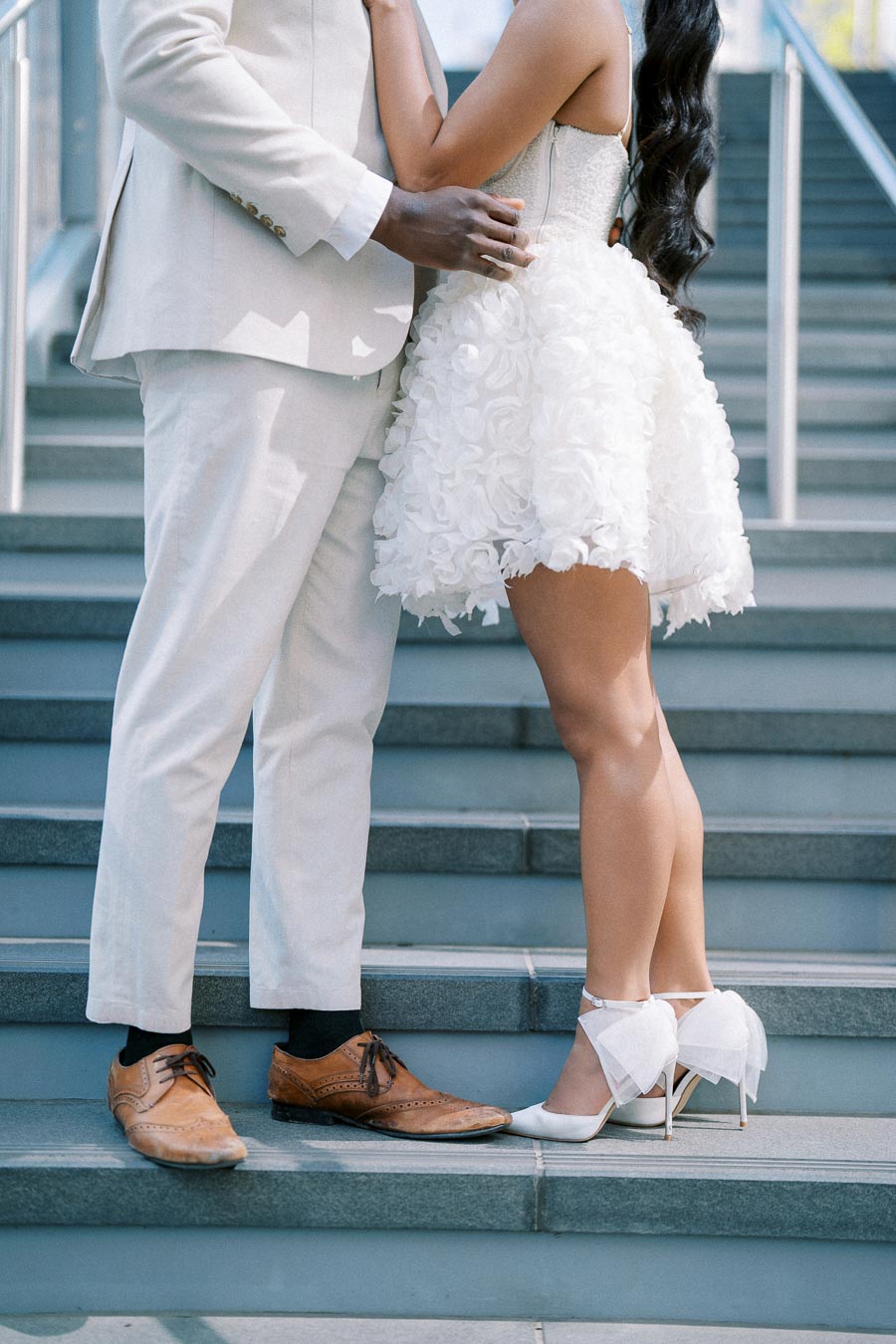 Elegant couple in fashionable attire standing on stairs, showcasing stylish brown shoes and white high heels with a floral dress, perfect for a modern wedding or engagement theme.