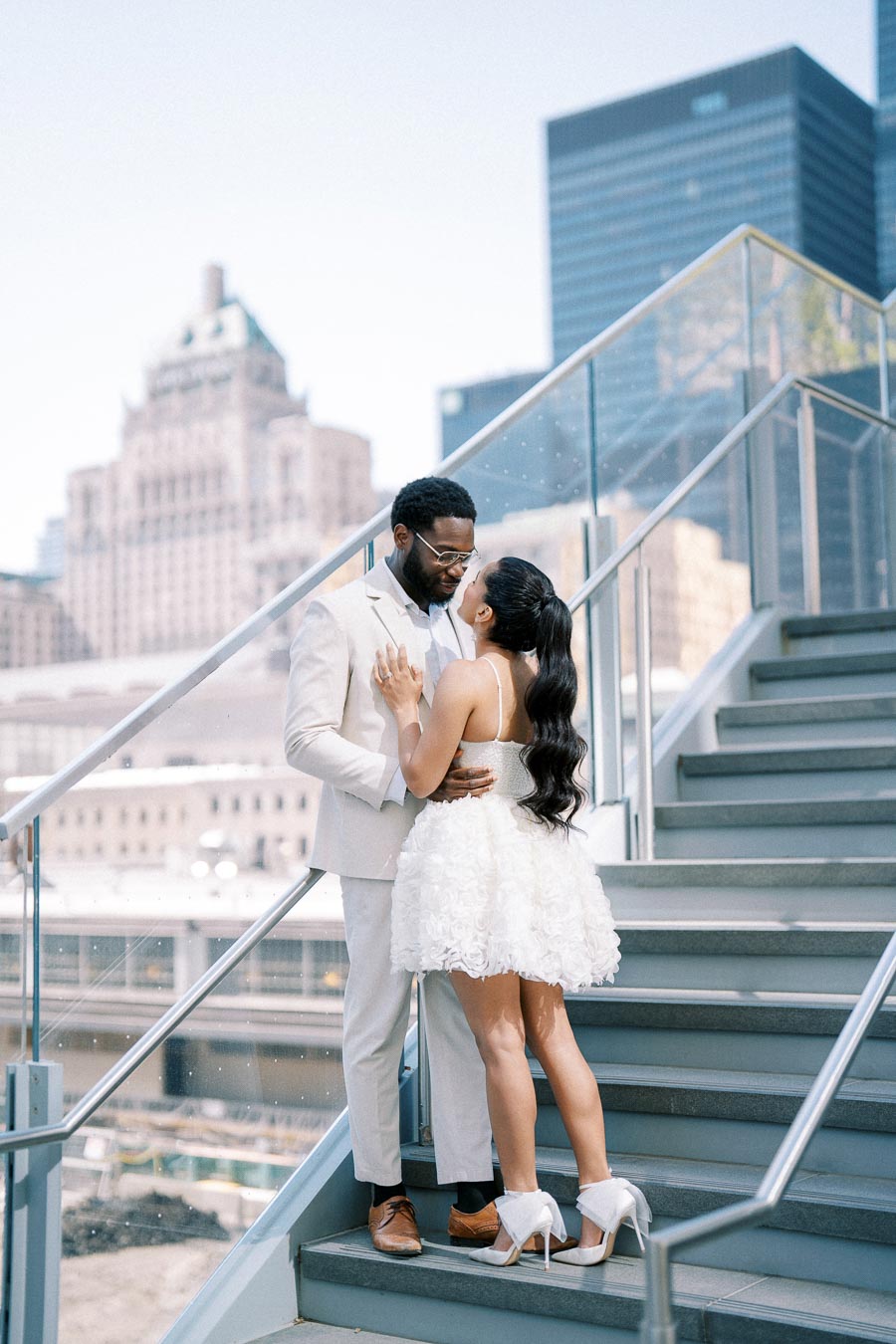 A couple in elegant white attire embraces on a modern outdoor staircase, set against a backdrop of city skyscrapers and a clear blue sky.