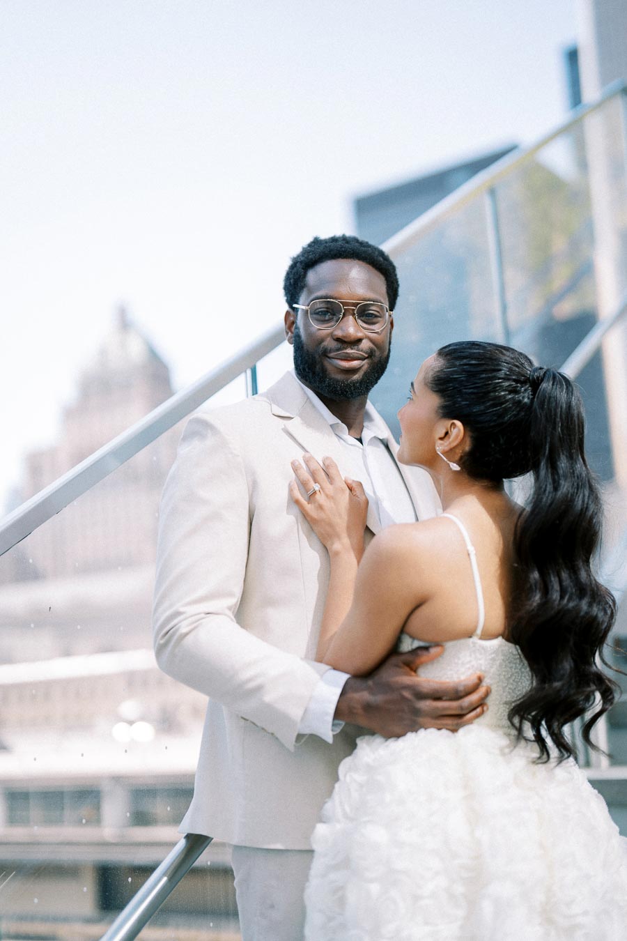 A couple in elegant attire standing on an outdoor staircase, with a cityscape backdrop, sharing a joyful moment. The man is wearing a light-colored suit and glasses, while the woman is in a white, textured dress. Bright and sunny day emphasizes their happiness and connection.