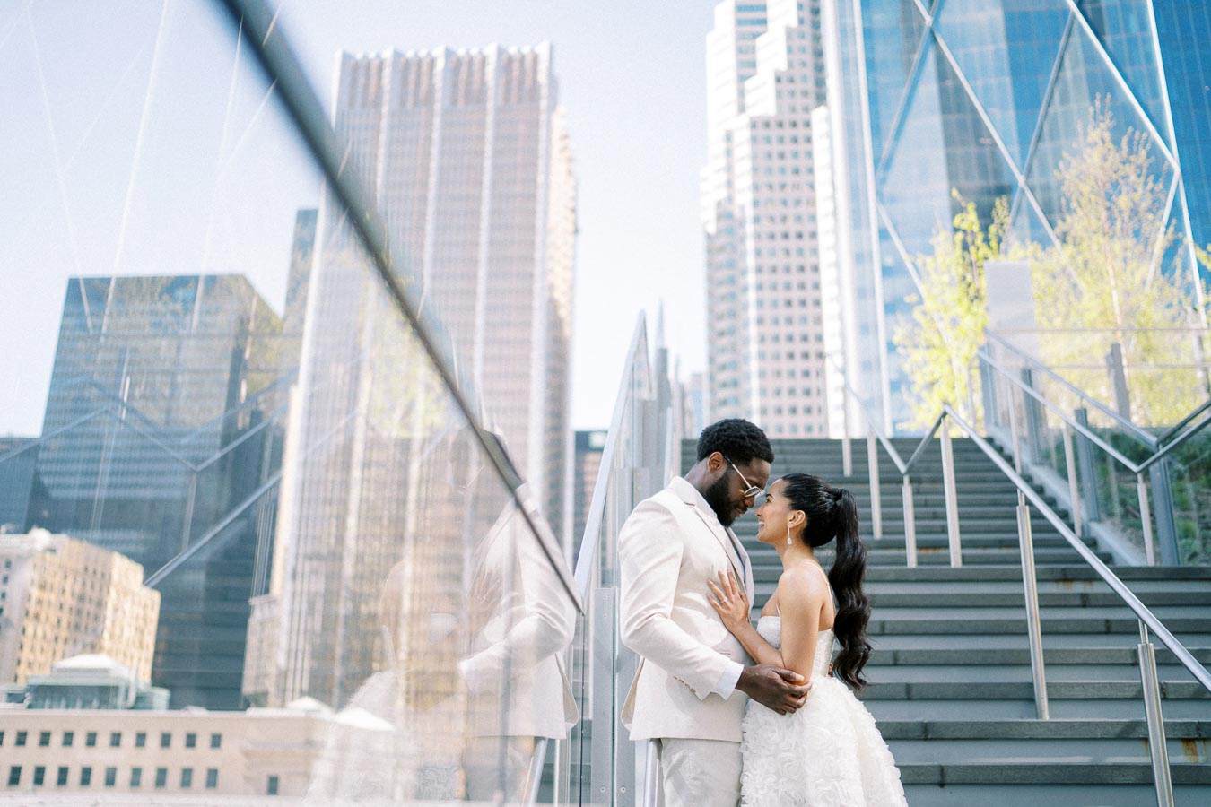 A couple in elegant attire embraces on a staircase with a city skyline in the background, surrounded by modern skyscrapers and clear blue sky.