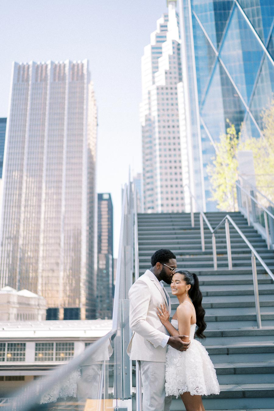 Couple embracing on urban staircase with skyscrapers in the background, dressed elegantly in white, capturing a modern and romantic moment amidst city architecture.