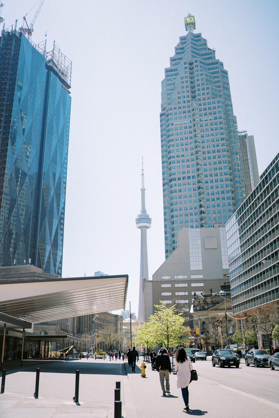 A bustling Toronto street scene featuring the iconic CN Tower, flanked by modern skyscrapers and pedestrians enjoying a sunny day.
