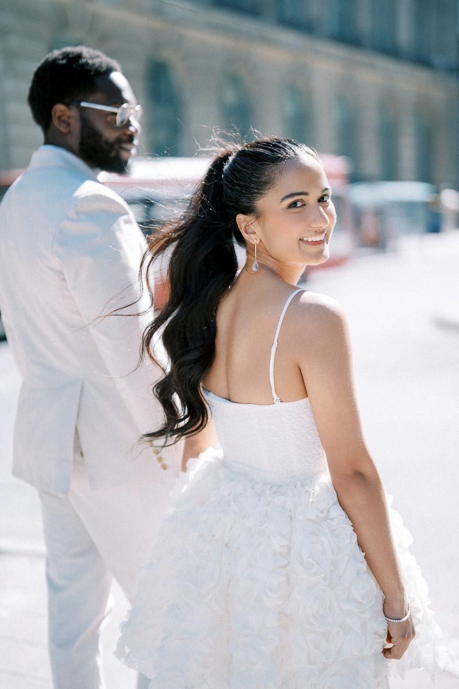 A couple elegantly dressed in white attire, walking outside with a cityscape backdrop. The woman is smiling over her shoulder in a white floral dress, while the man wears a white suit and sunglasses.