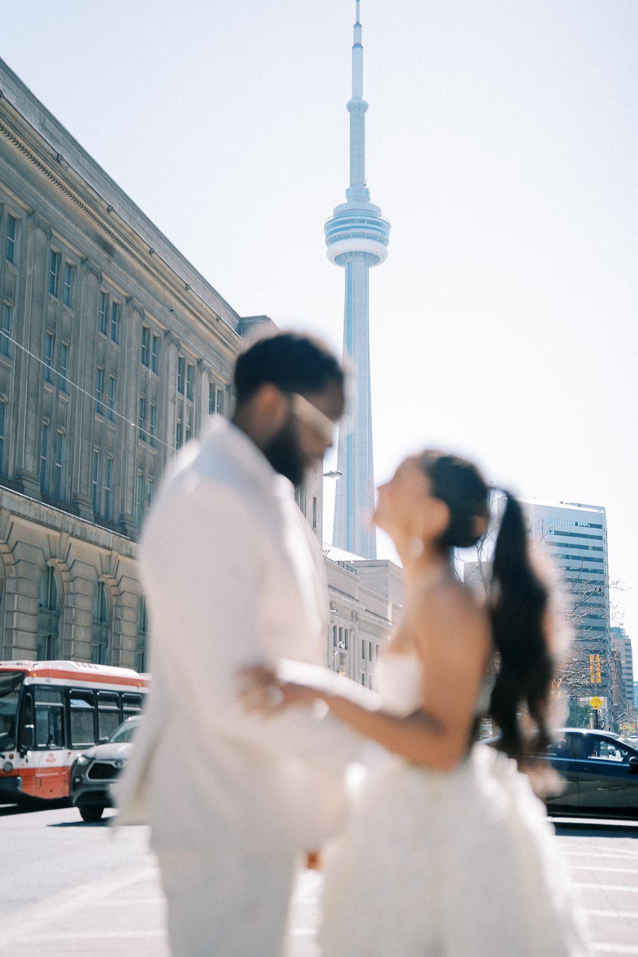 A couple in wedding attire standing in front of the iconic CN Tower in Toronto, with a city street and historic building in the background.
