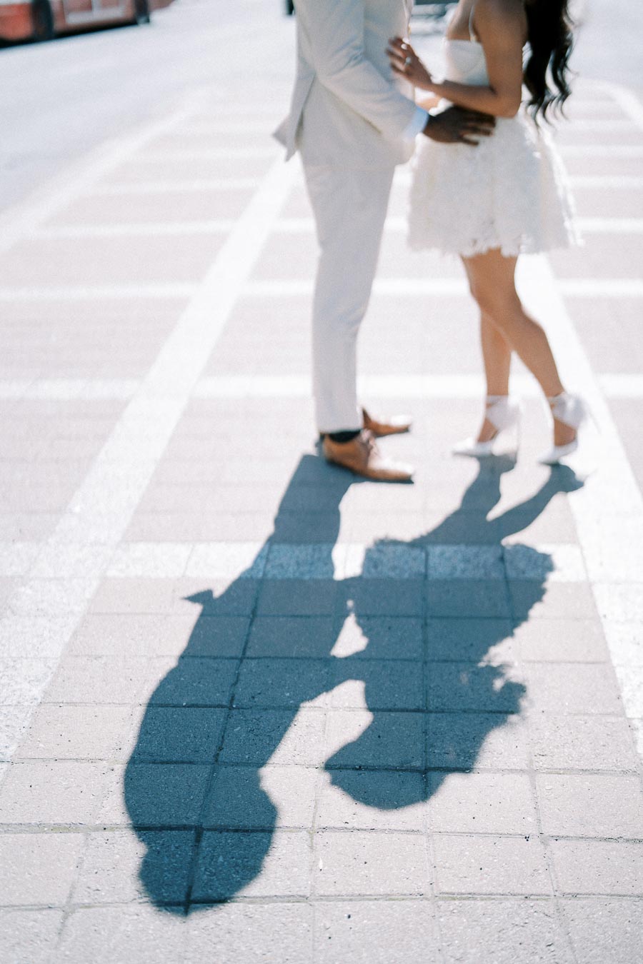 A couple in elegant attire embraces on a sunny sidewalk, their shadows cast prominently on the pavement.