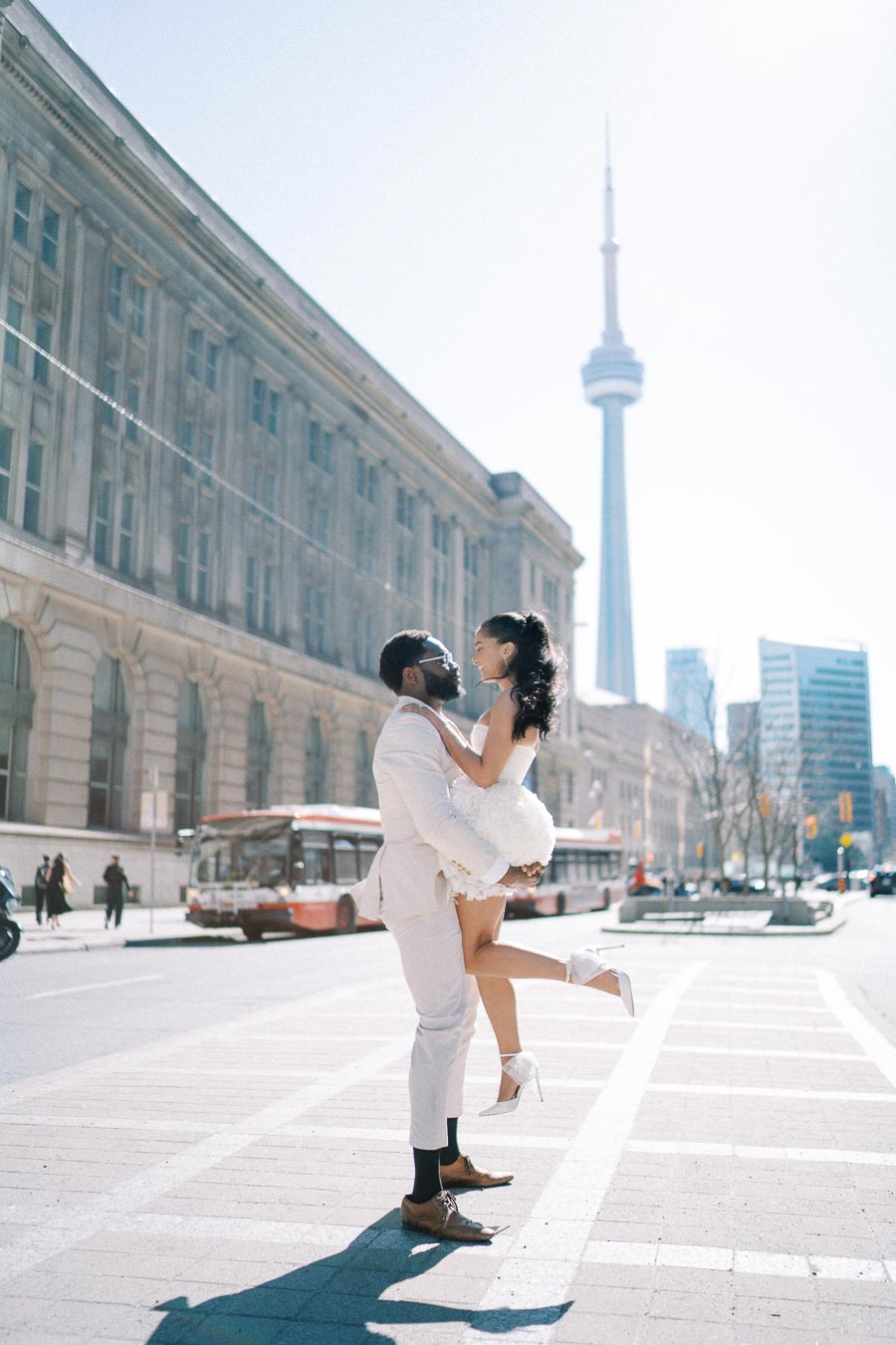 A romantic couple joyfully embracing on a city street with the CN Tower in the background, capturing a picturesque moment in Toronto.