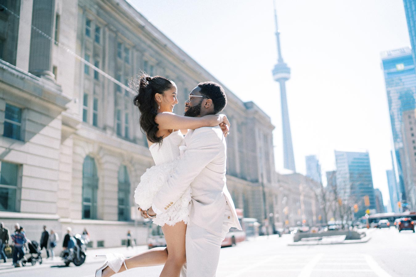 A happy couple in white wedding attire joyfully embrace on a city street with the iconic CN Tower in the background, capturing a moment of love in an urban setting.