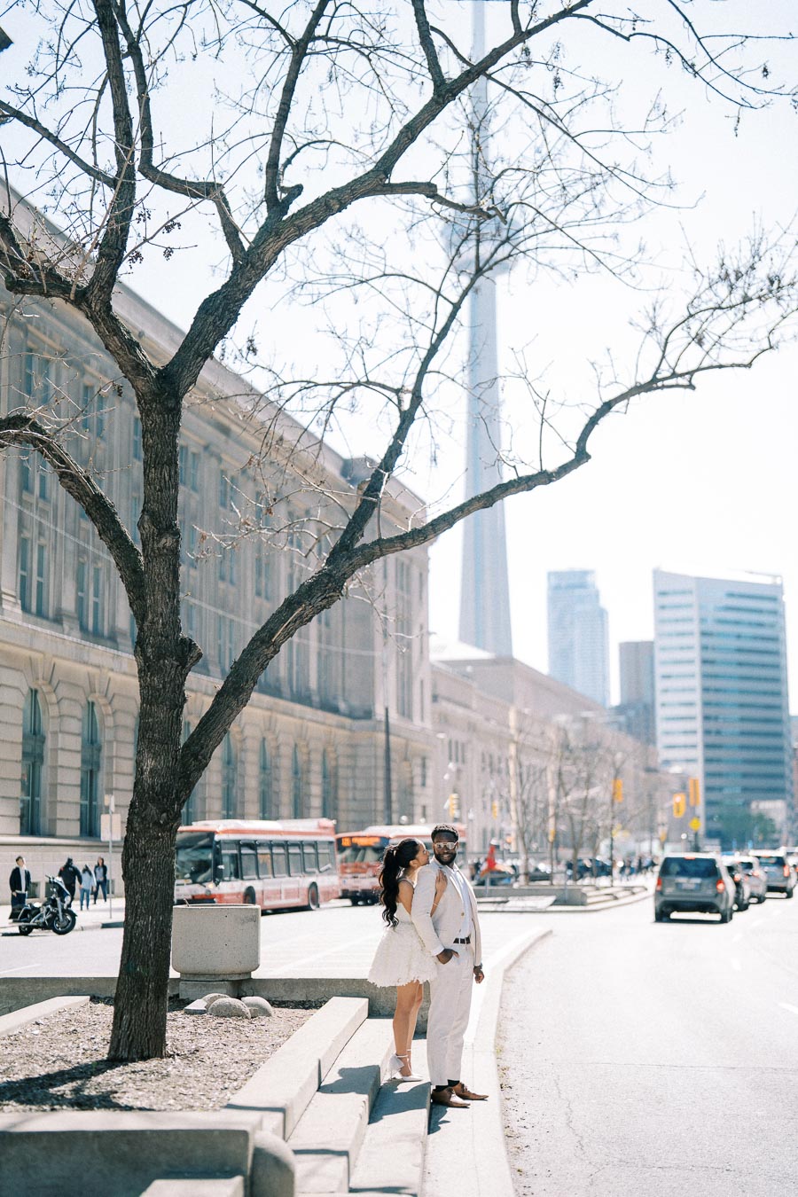 A couple in stylish white attire standing on a city street with a towering landmark and modern buildings in the background, capturing an urban and elegant vibe.