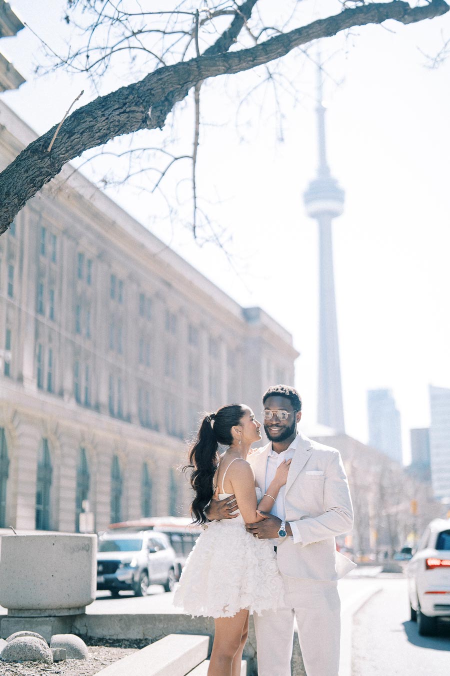 Couple embracing on a city street with iconic tower in the background, sunlight filtering through bare tree branches.