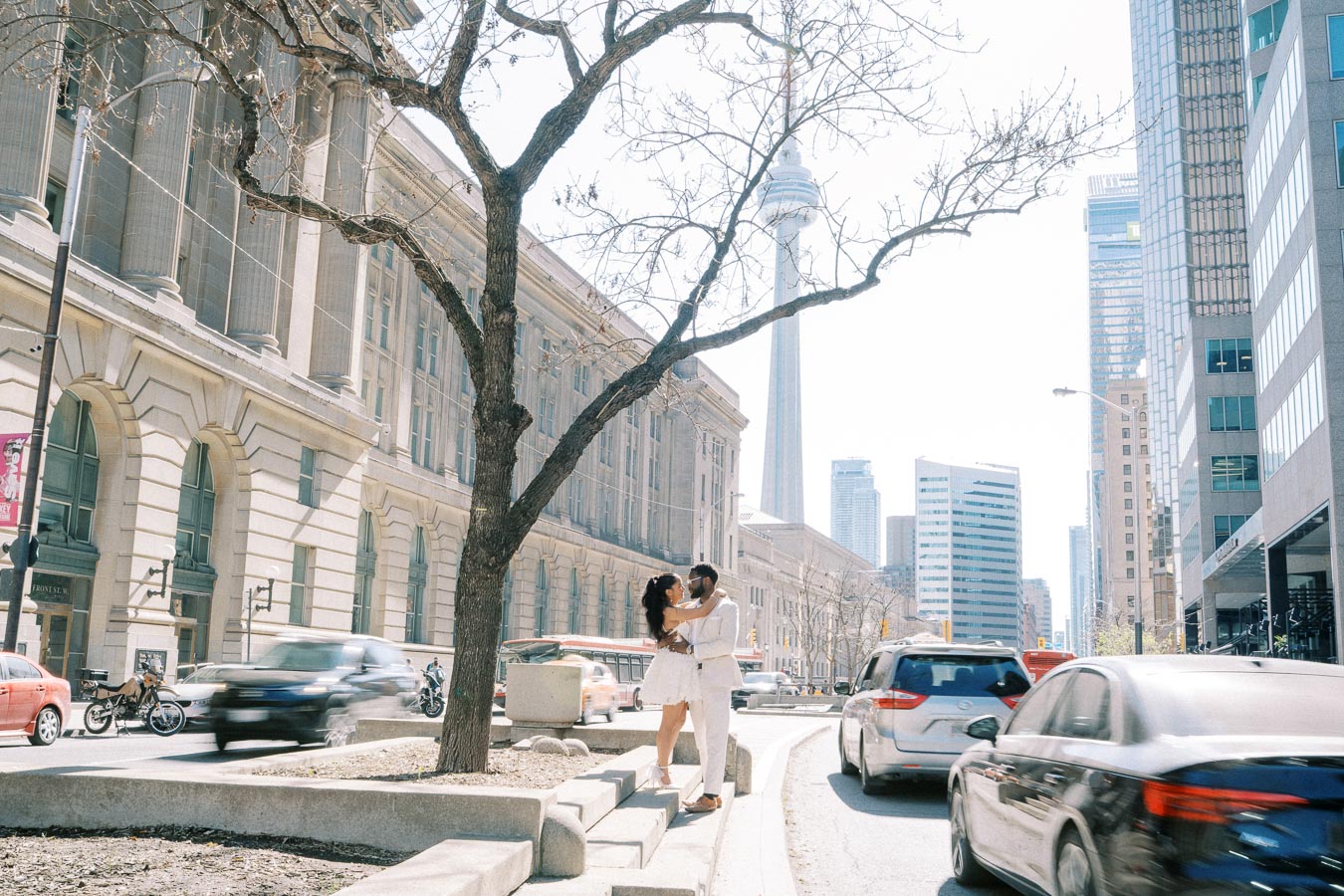 A couple embracing on a busy urban street with tall buildings in the background, including a prominent tower under a clear sky.