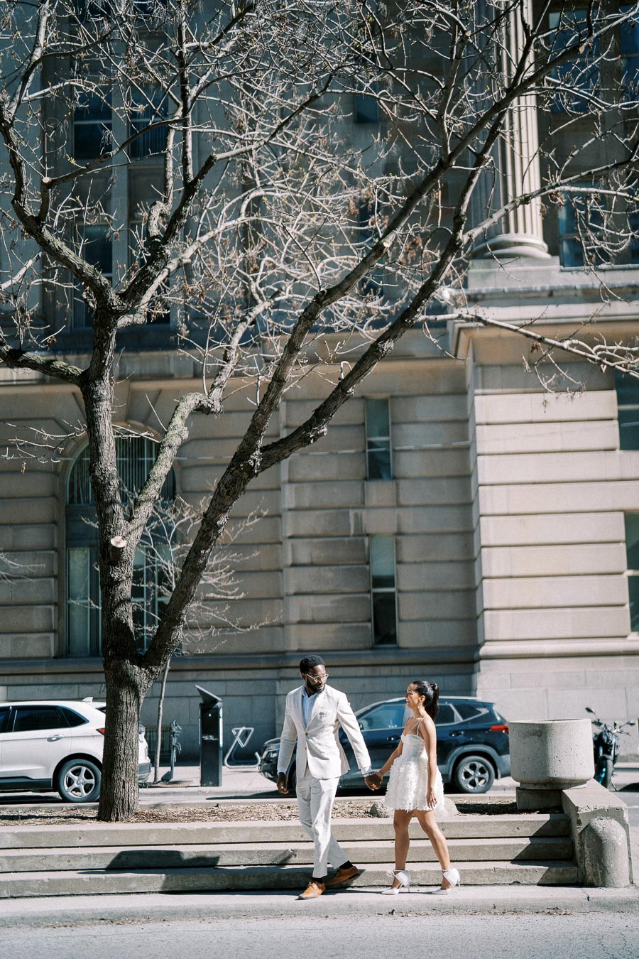 A couple in stylish attire holding hands walks past a historic building with bare trees and parked cars.